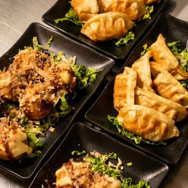 Plates of takoyaki with bonito flakes and gyoza with dipping sauce on a metal surface.