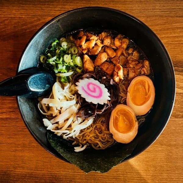 Overhead view of a black bowl of ramen with chicken, eggs, scallions, and a pink swirl.